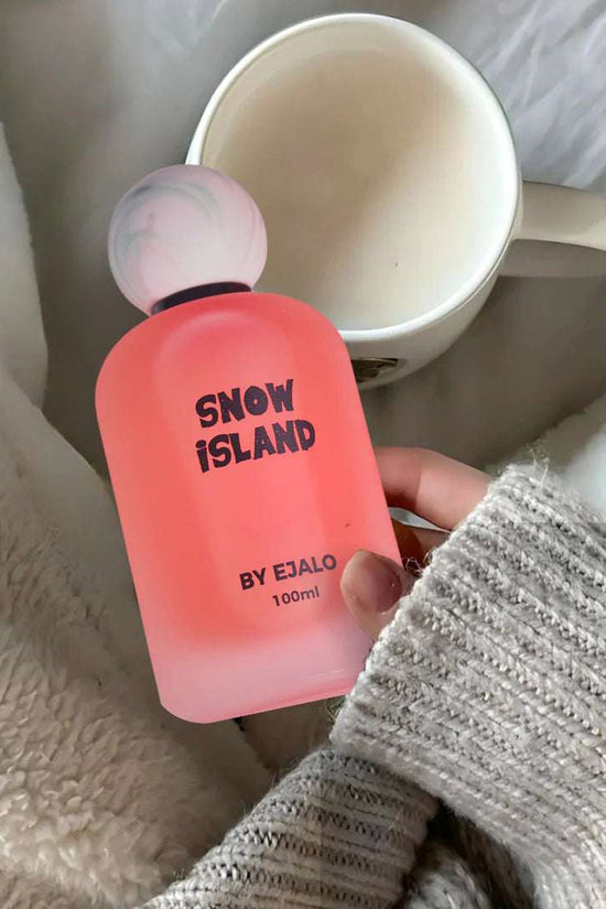 Snow Island perfume 100ml presented in a pink frosted bottle, featuring a round cap, beside a coffee mug and cozy blanket