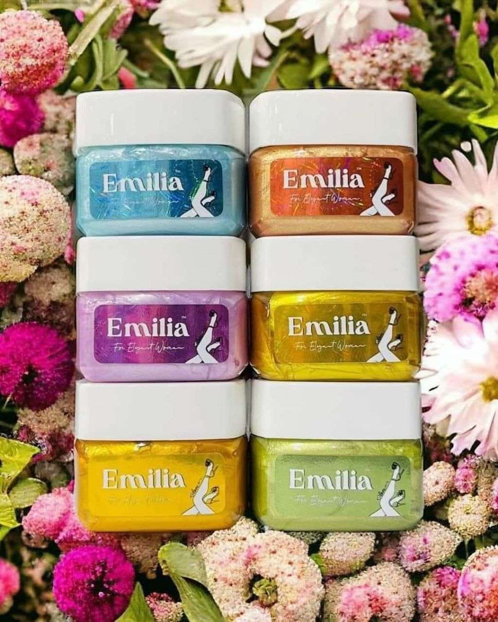 Collection of Emilia body perfumes in colorful jars arranged among flowers, featuring Emilia High Light body perfume