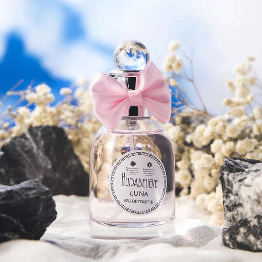 Huda Believe perfume in an elegant bottle with a pink bow, surrounded by natural stones and flowers, perfect for alluring fragrance lovers.